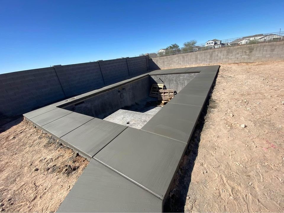 The image shows a partially constructed rectangular swimming pool with concrete walls, while the concrete pool deck installation is still underway. Wooden pallets are inside the pool, and a concrete wall encloses the site. The sky is clear and blue, offering perfect weather for continuing construction.