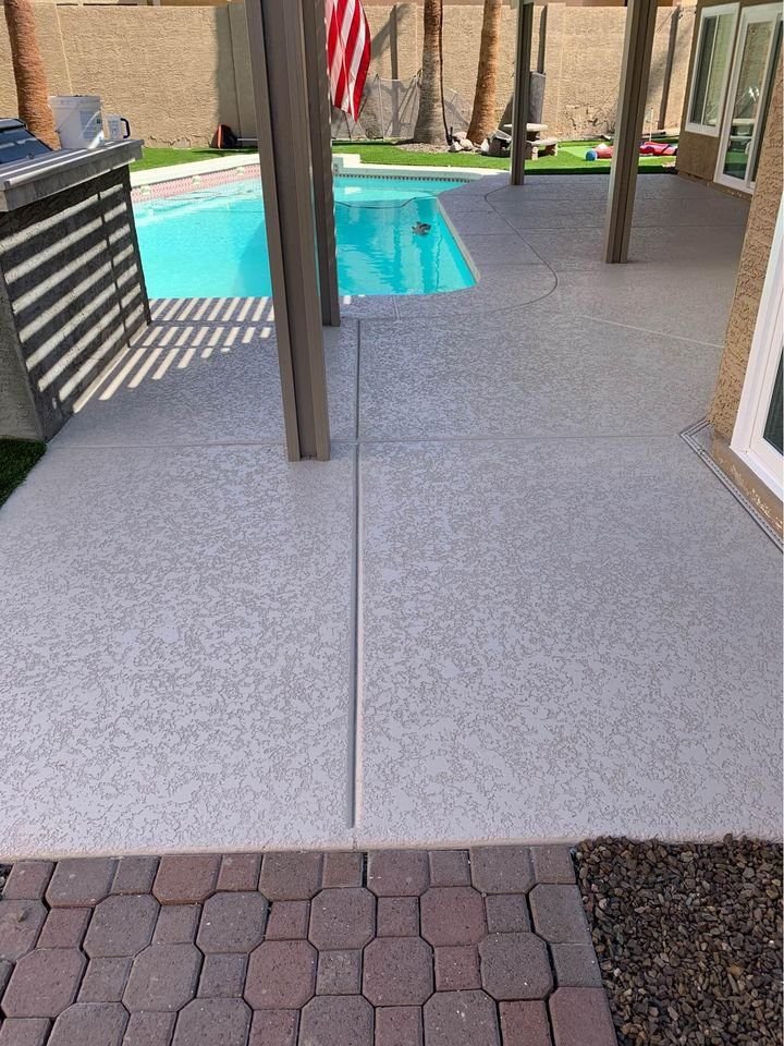 A backyard pool area features a textured, light-colored concrete patio resembling a Phoenix concrete pool deck and a red brick pathway. There's a striped pool house on the left, a grass patch, and an American flag beside the water. The pool's edge casts a shadow on the patio.