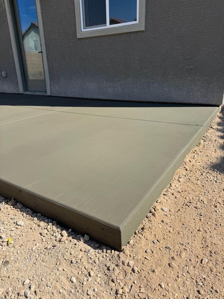 A freshly poured, smooth concrete patio in Phoenix, AZ, lies adjacent to a house with light-colored walls, a window, and a door visible. The ground around the patio is rough and unfinished, consisting of gravel.